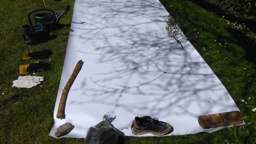 Residuals 3, Dartington Hall Gardens on Saturday 20 April 2024. Shadows on the surface of the drawing paper prior to the performance. This photograph prior to the performance, The drawing paper - measuring about 4m by 1.5m - set on the grass with tools and watering cans beside it.